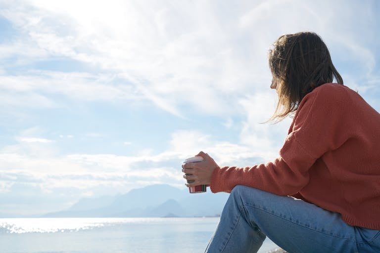 Introvert OCD. A woman in casual attire enjoys a hot drink while gazing at the sea, epitomizing relaxation.