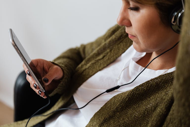 A woman sitting indoors, using a smartphone and headphones to listen to music.