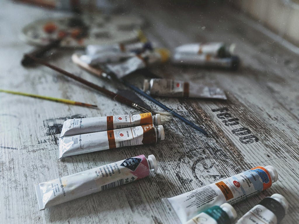 Close-up of various acrylic paint tubes and brushes on a worn, textured wooden surface.