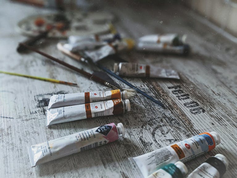 Close-up of various acrylic paint tubes and brushes on a worn, textured wooden surface.