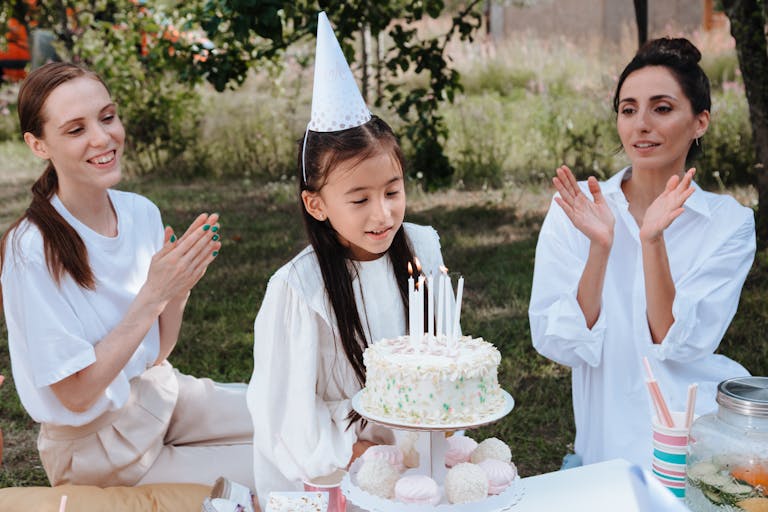 Family celebrates a young girl's birthday outdoors with cake and clapping.