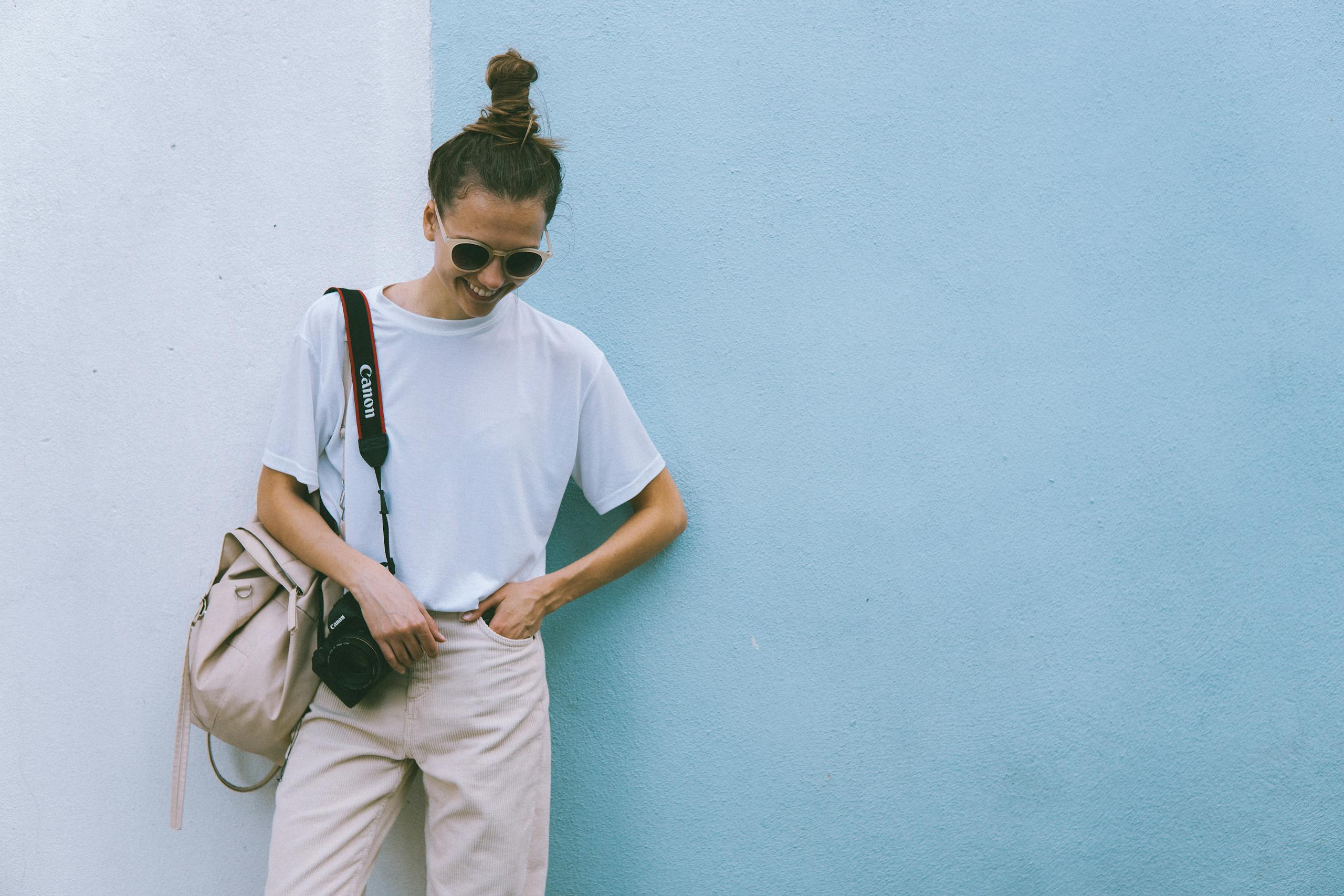 Stylish woman posing with camera in casual attire against a blue wall.