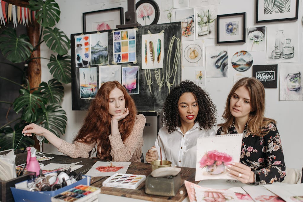 Three women in an art studio discuss watercolor paintings, surrounded by artwork and painting materials.