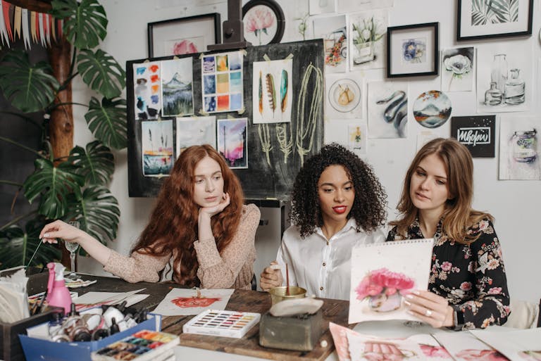 Three women in an art studio discuss watercolor paintings, surrounded by artwork and painting materials.