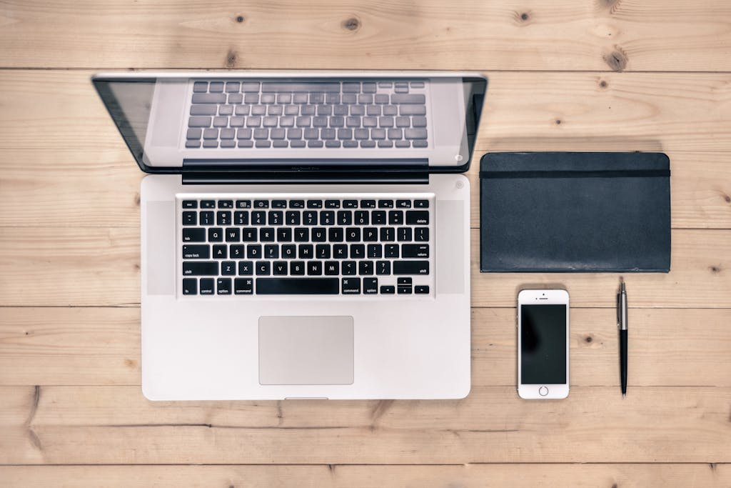 Top view of a tidy workspace featuring a laptop, smartphone, notebook, and pen on a wooden desk.