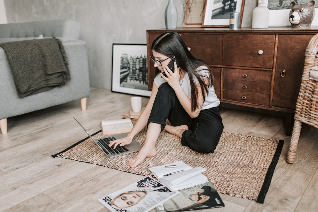 Young woman working from home, balancing laptop and phone calls on the floor.
