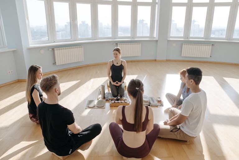 A diverse group of adults meditating in a spacious, sunlit studio with city views.