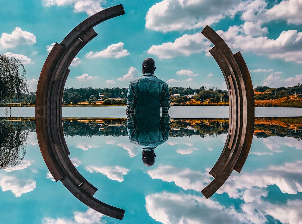 Introvert Autism Support. A man in a denim jacket sits by abstract sculpture reflecting on the water under a blue sky.