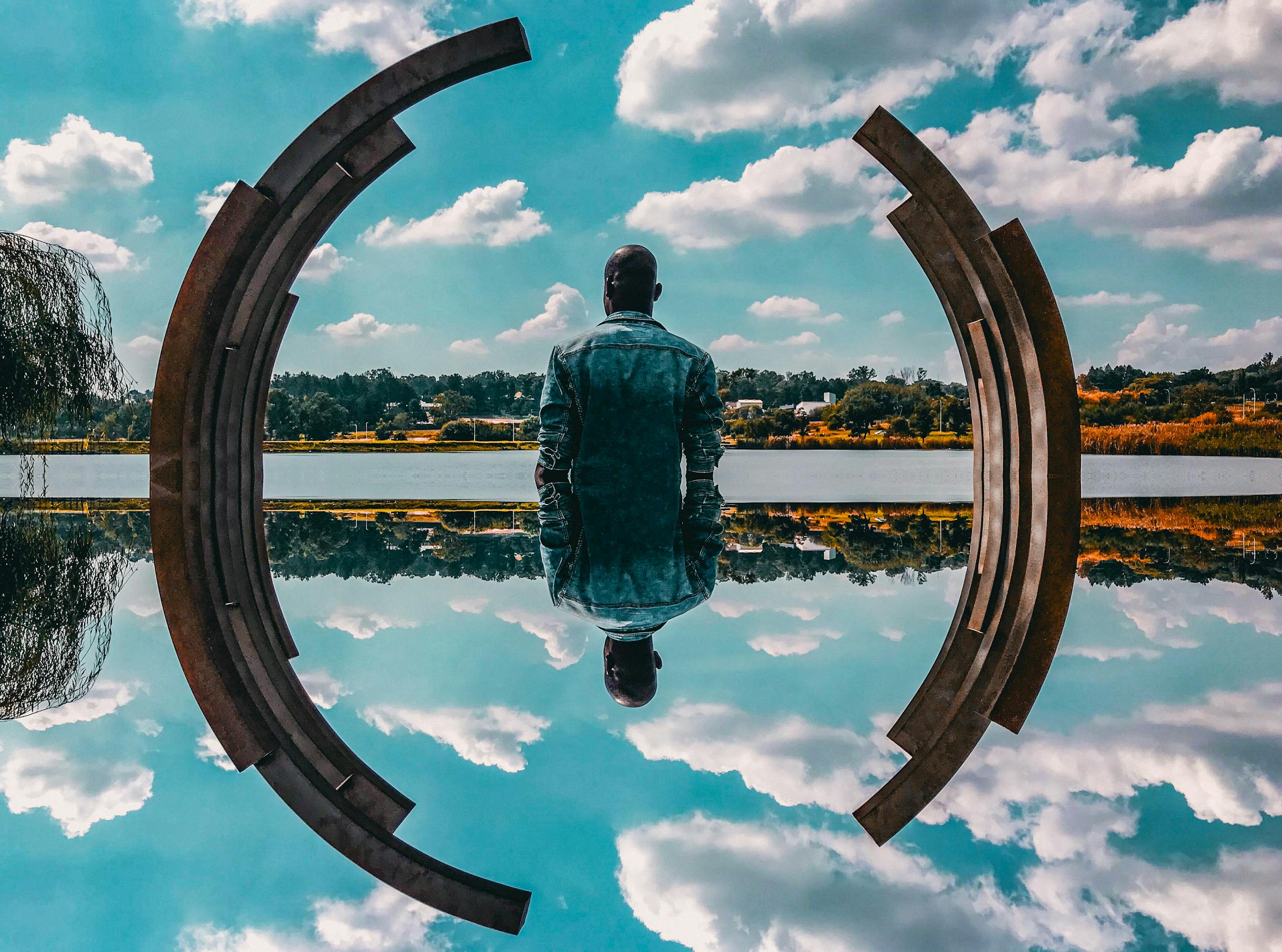 A man in a denim jacket sits by abstract sculpture reflecting on the water under a blue sky.