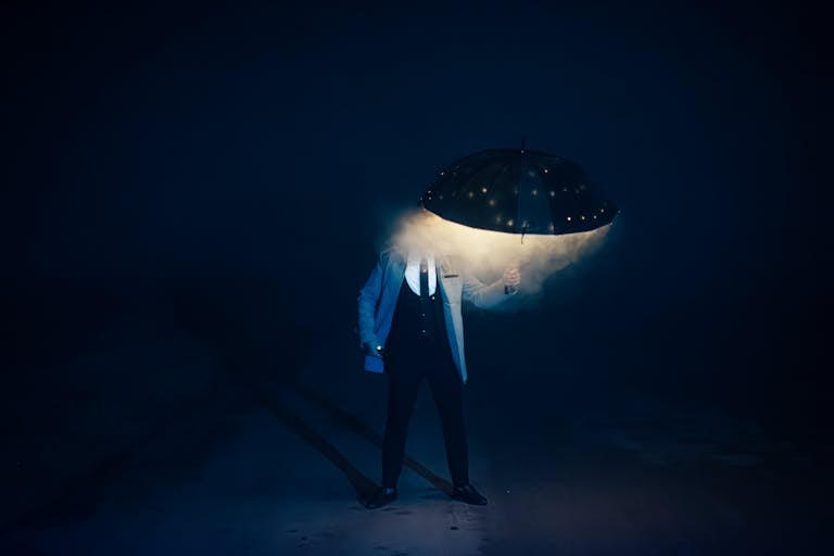 A person in a suit stands in foggy darkness holding an LED-lit umbrella, creating a mysterious scene.