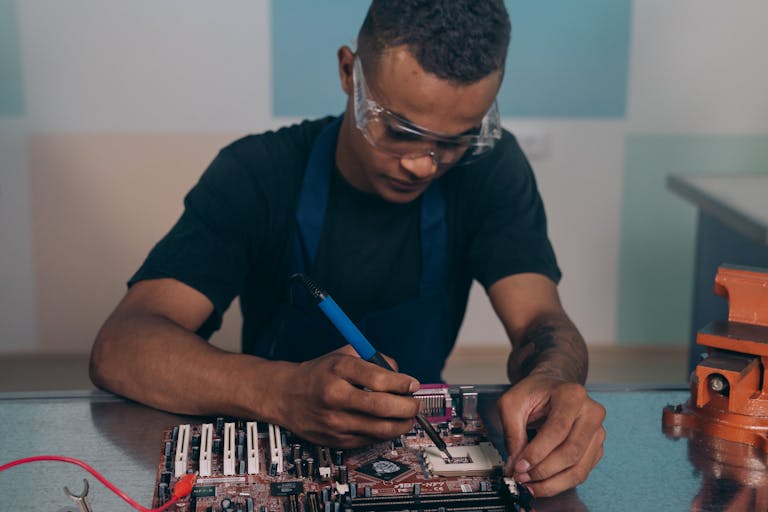 A technician wearing safety glasses works intently on a computer motherboard with a soldering iron.