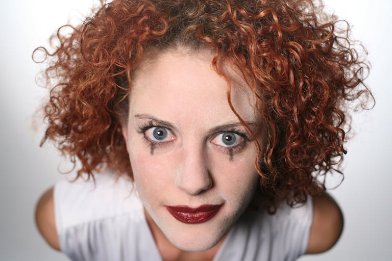 A woman with curly red hair and dramatic clown makeup looking upwards, striking a bold pose.