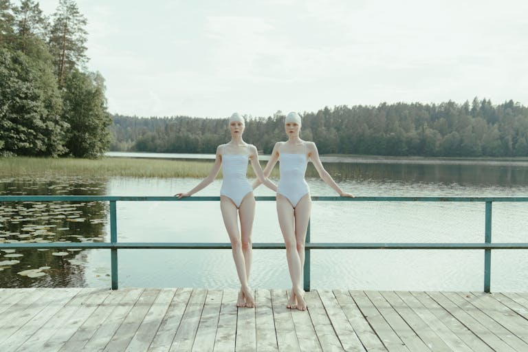 Two women in white swimsuits standing on a dock at a serene lake surrounded by trees.