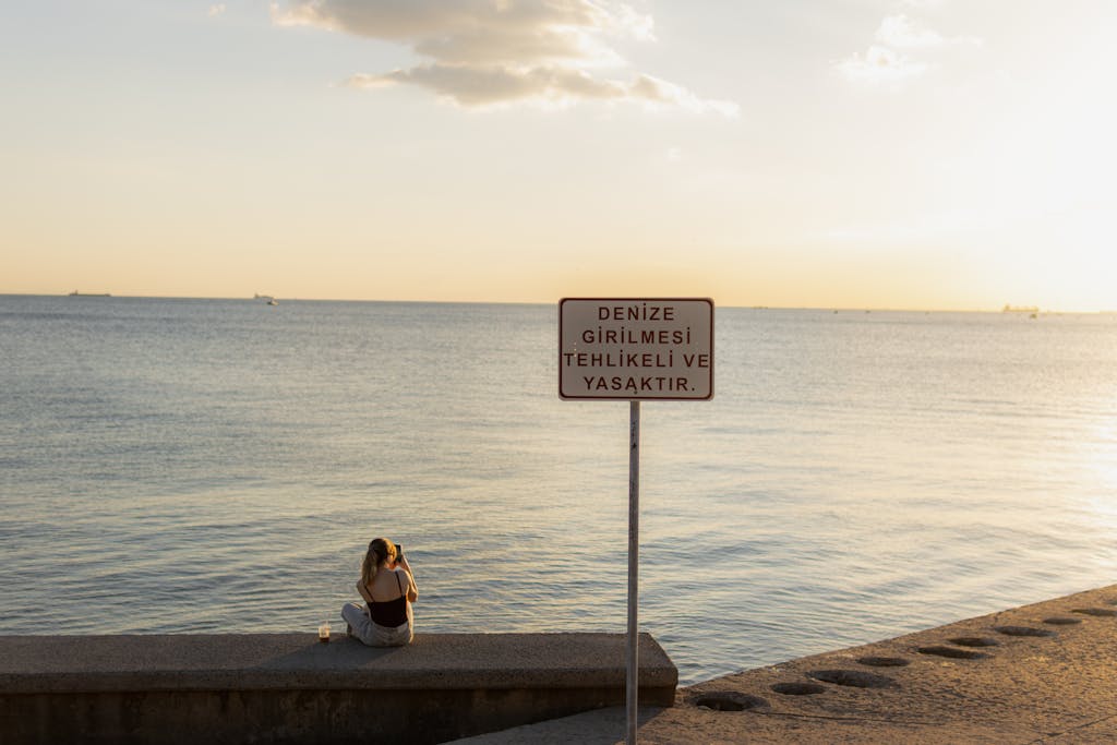 Woman sits near the sea at sunset in Istanbul, with a sign warning not to enter the water.