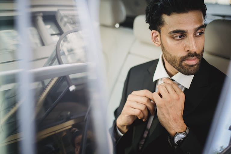 A bearded man in a black suit sitting inside a car, adjusting his tie and looking serious.