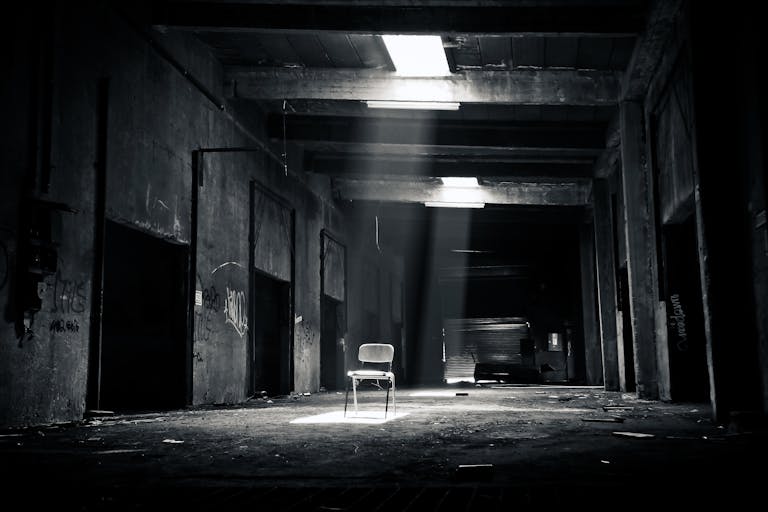A haunting black and white photo of an abandoned warehouse with a solitary chair illuminated by sunlight through a window.