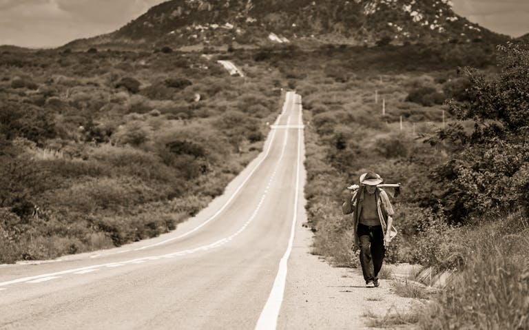A solitary man walks along a long, scenic road in rural Brazil, under a vast sky.
