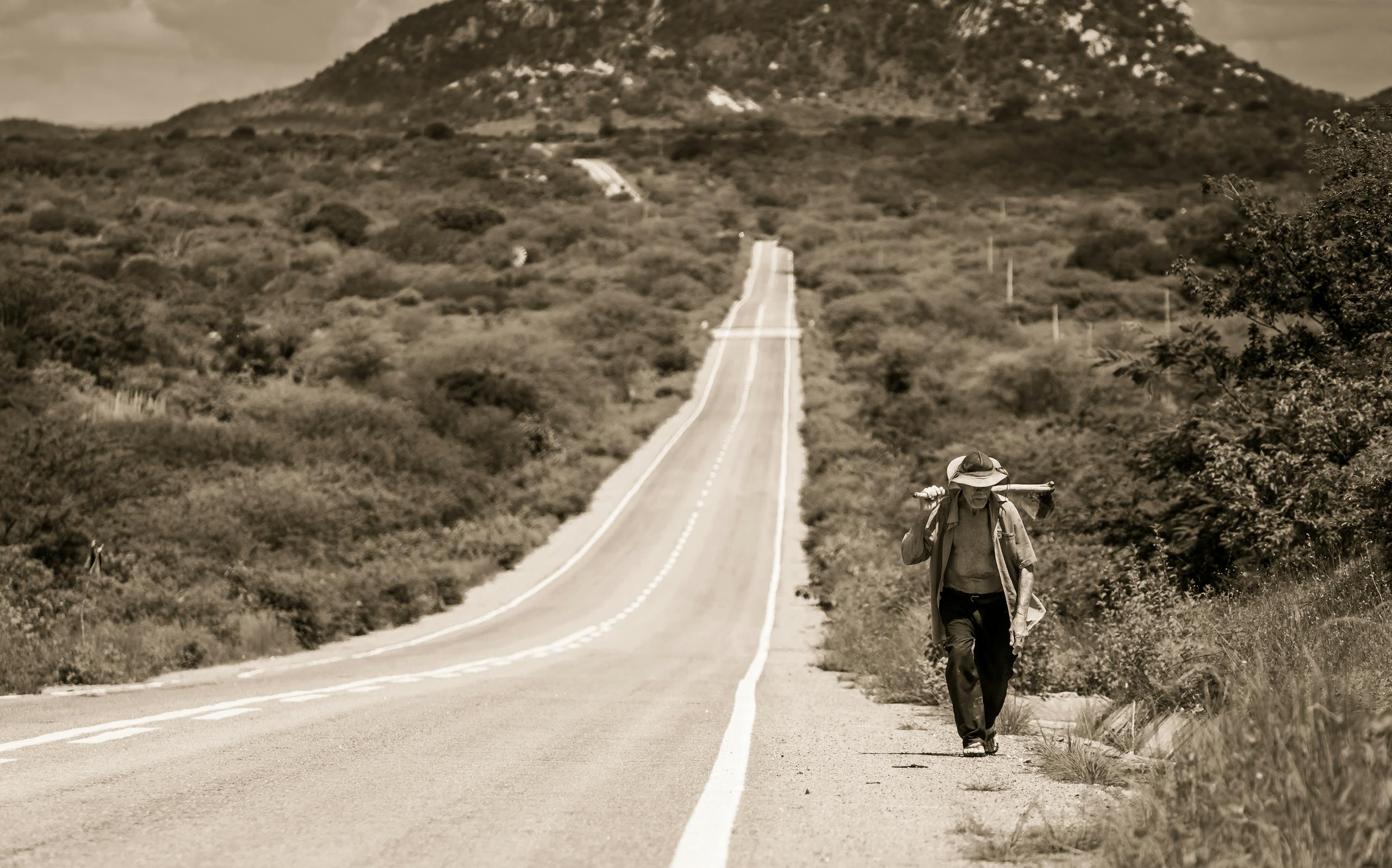 A solitary man walks along a long, scenic road in rural Brazil, under a vast sky.
