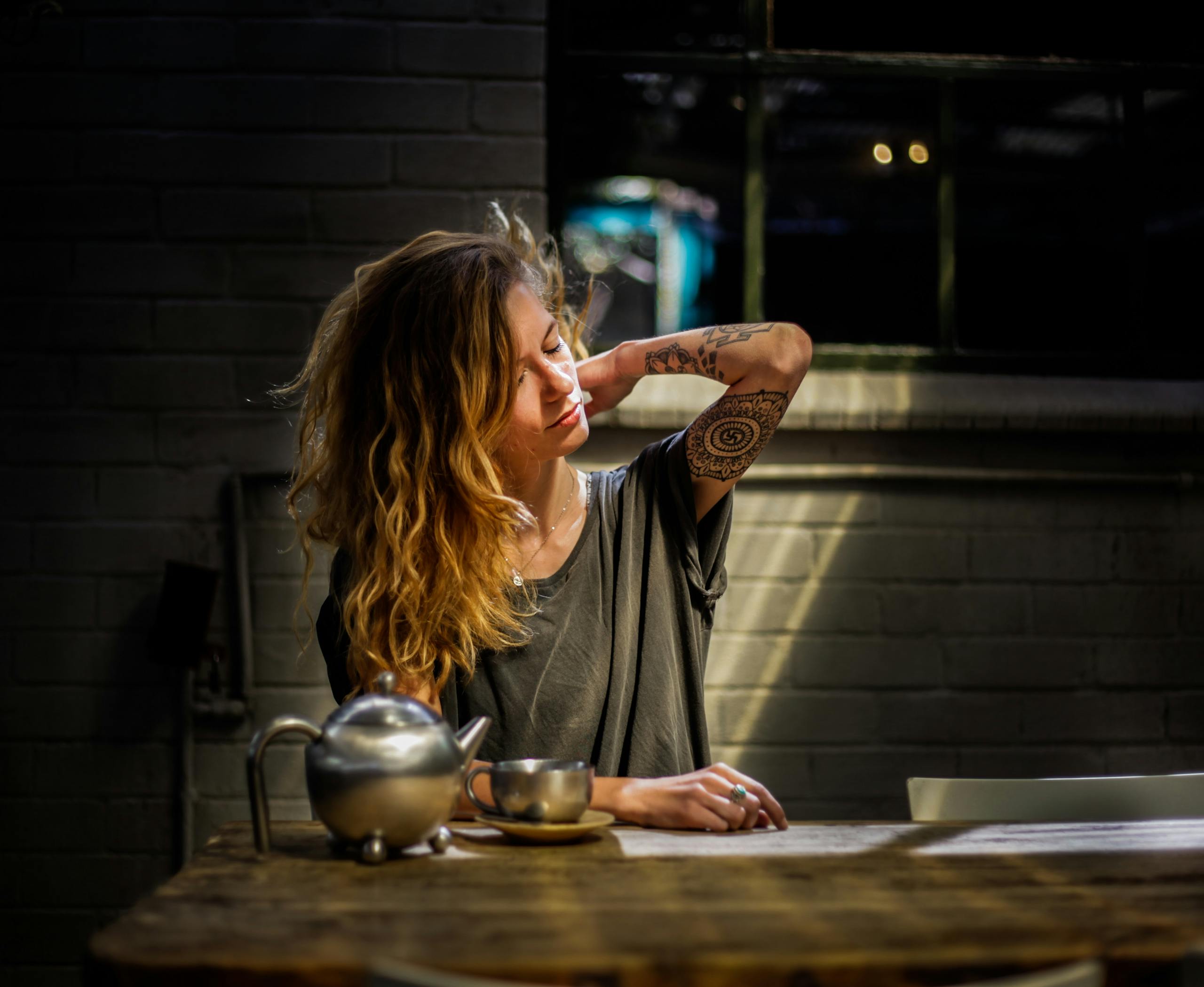 A woman with tattoos relaxes with a cup of tea in a sunlit room. Casual and serene moment.