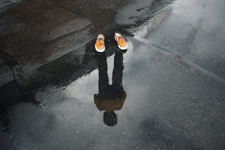 Reflection of legs and shoes in a puddle on a wet street, creating a surreal urban scene.