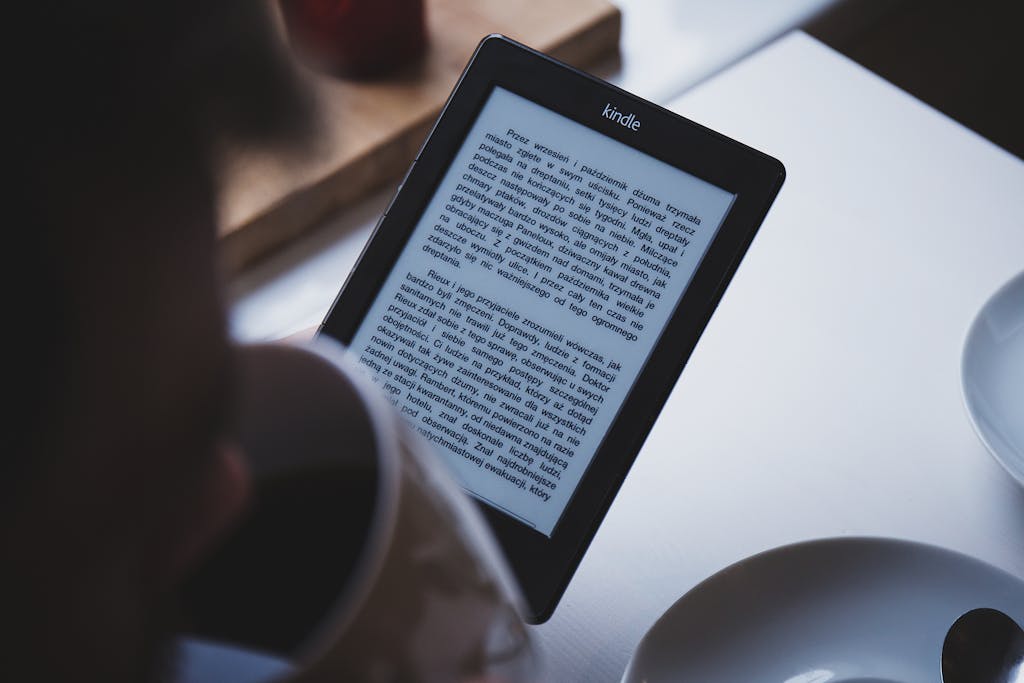 A person reads on a Kindle e-reader while holding a cup of coffee, indoors.