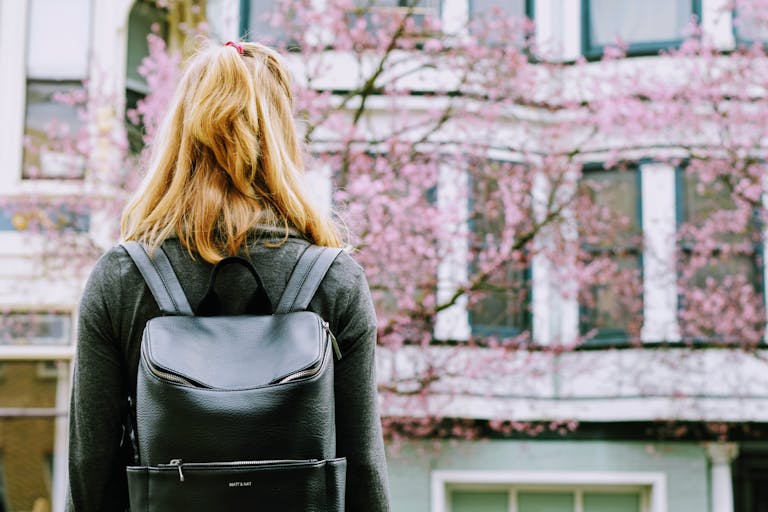 Blonde woman with a backpack stands by urban cherry blossom, hinting at travel and fashion.