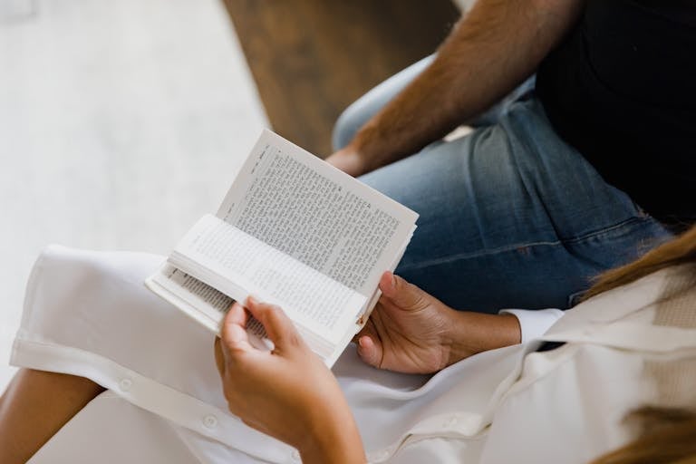 Two people reading a book together, one wearing a white dress, indoors.