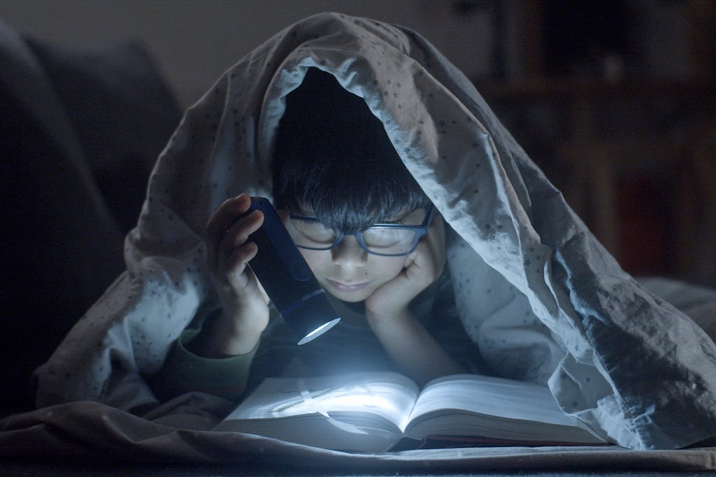 Young child enjoying a bedtime story under a blanket with a flashlight.