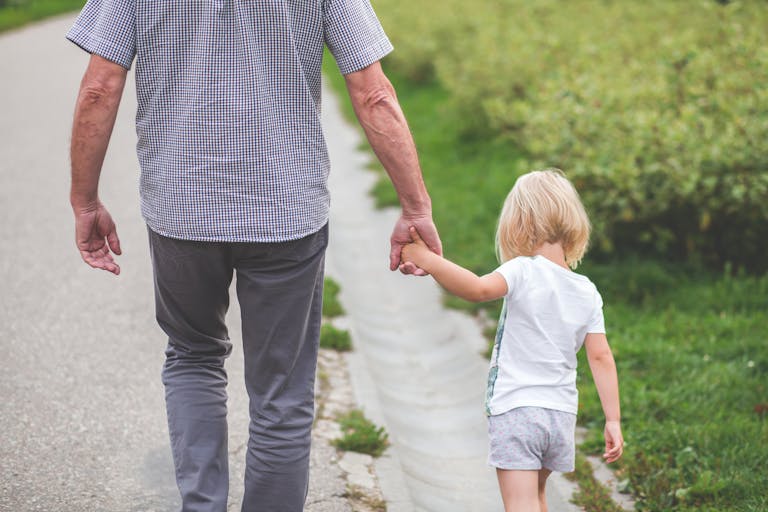 A grandparent holds hands with a child, walking on a path in a park, symbolizing family and togetherness.