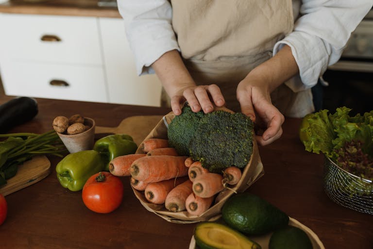 Hands arranging fresh broccoli and carrots with other vegetables on a kitchen counter.