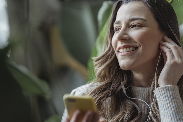 Happy young woman with long wavy hair smiling and adjusting earphones while listening to music via mobile phone