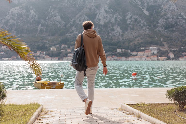Man in a hoodie walking on a dock towards calm waterfront with mountains in the background.