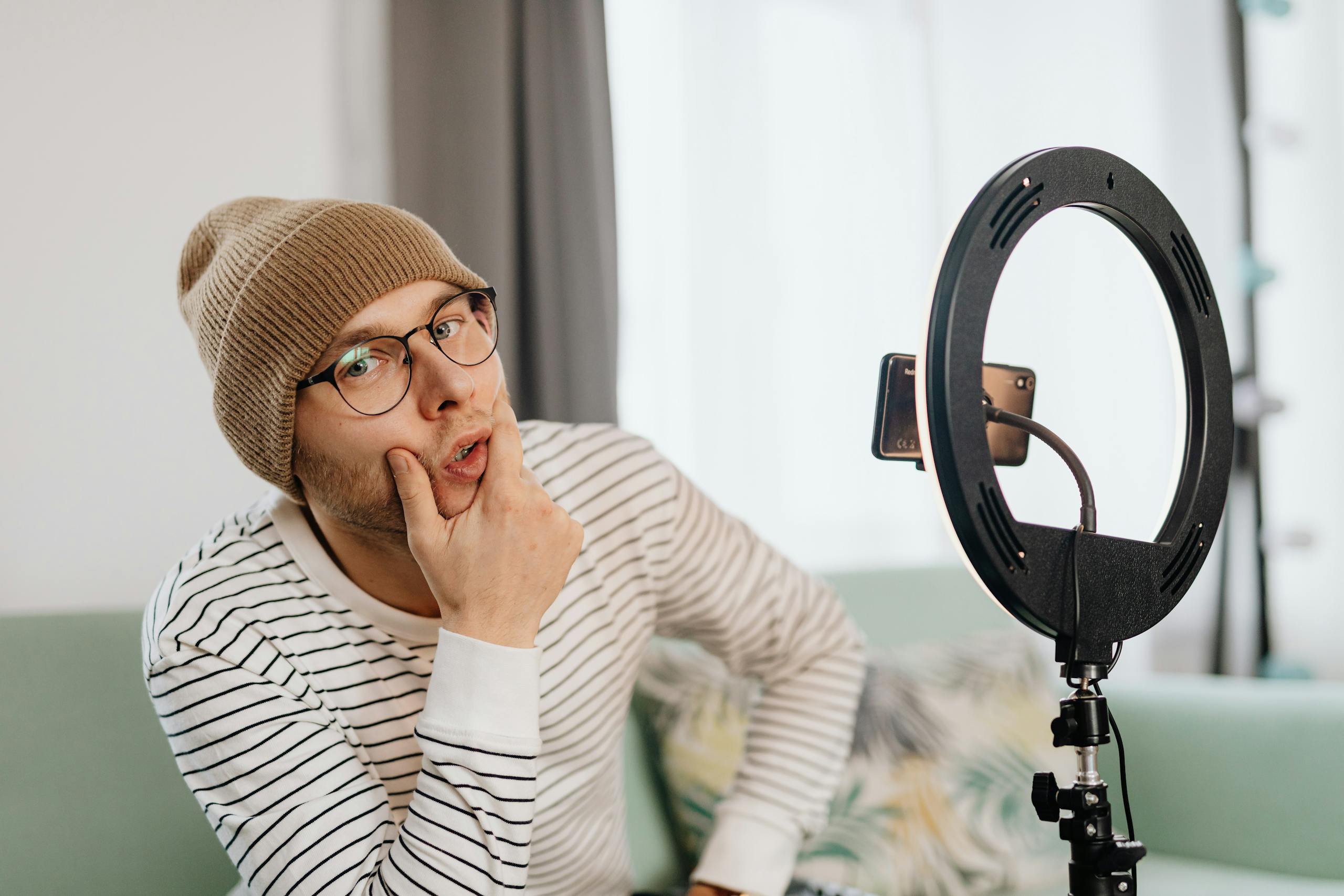 Young content creator with beanie and glasses posing in front of ring light indoors.