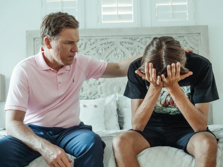 Compassionate father consoling upset teenage son on bed indoors