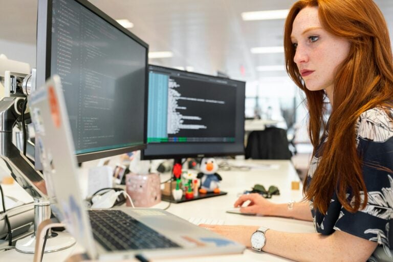 Focused female software engineer codes on dual monitors in modern office.