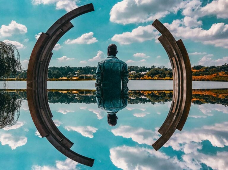 Man in denim jacket sitting contemplatively by abstract sculpture and water.