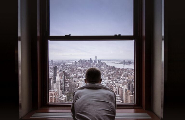Man reflecting while overlooking New York City skyline through window