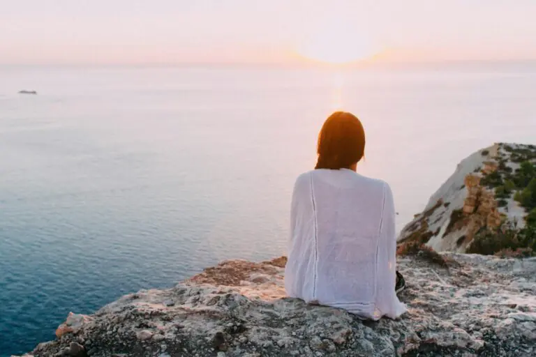 Woman sitting on rocky cliff gazing at ocean at serene sunset peacefully.