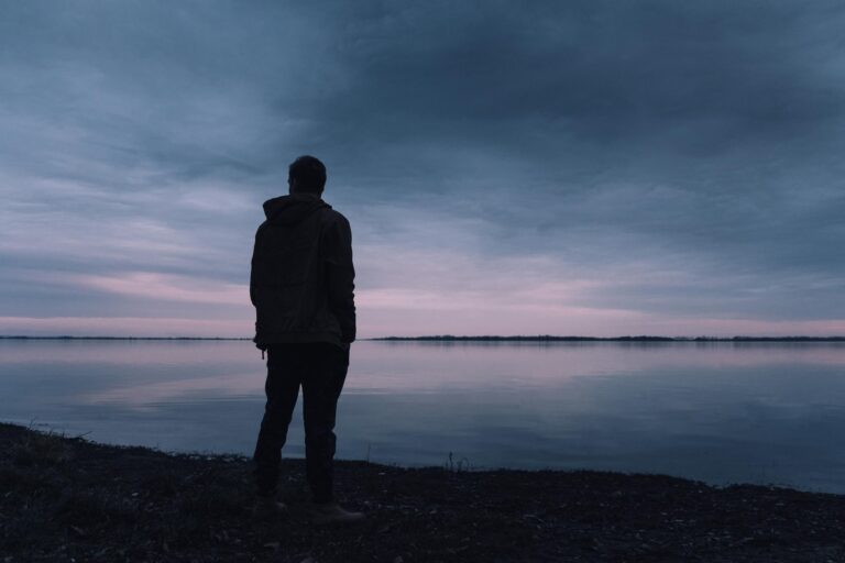 Silhouette of man in jacket gazing at lake at peaceful sunset.
