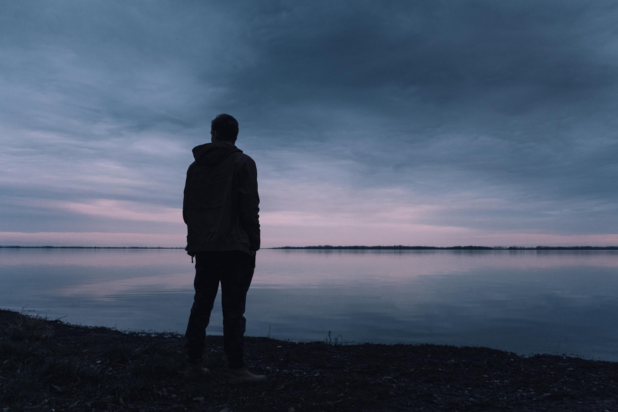Silhouette of man in jacket gazing at lake at peaceful sunset.