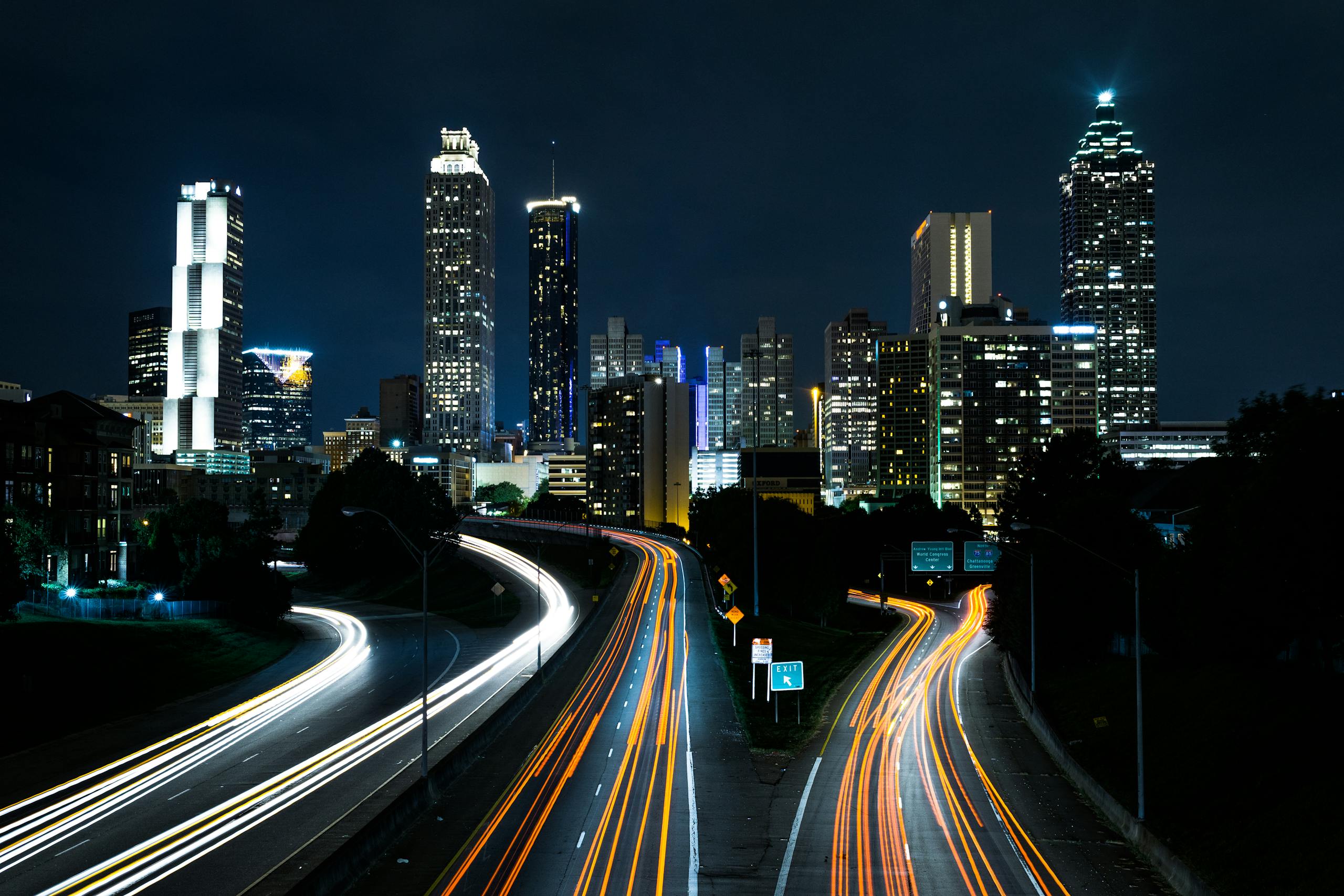 Vibrant cityscape at night with light trails on highway beneath towering skyscrapers.