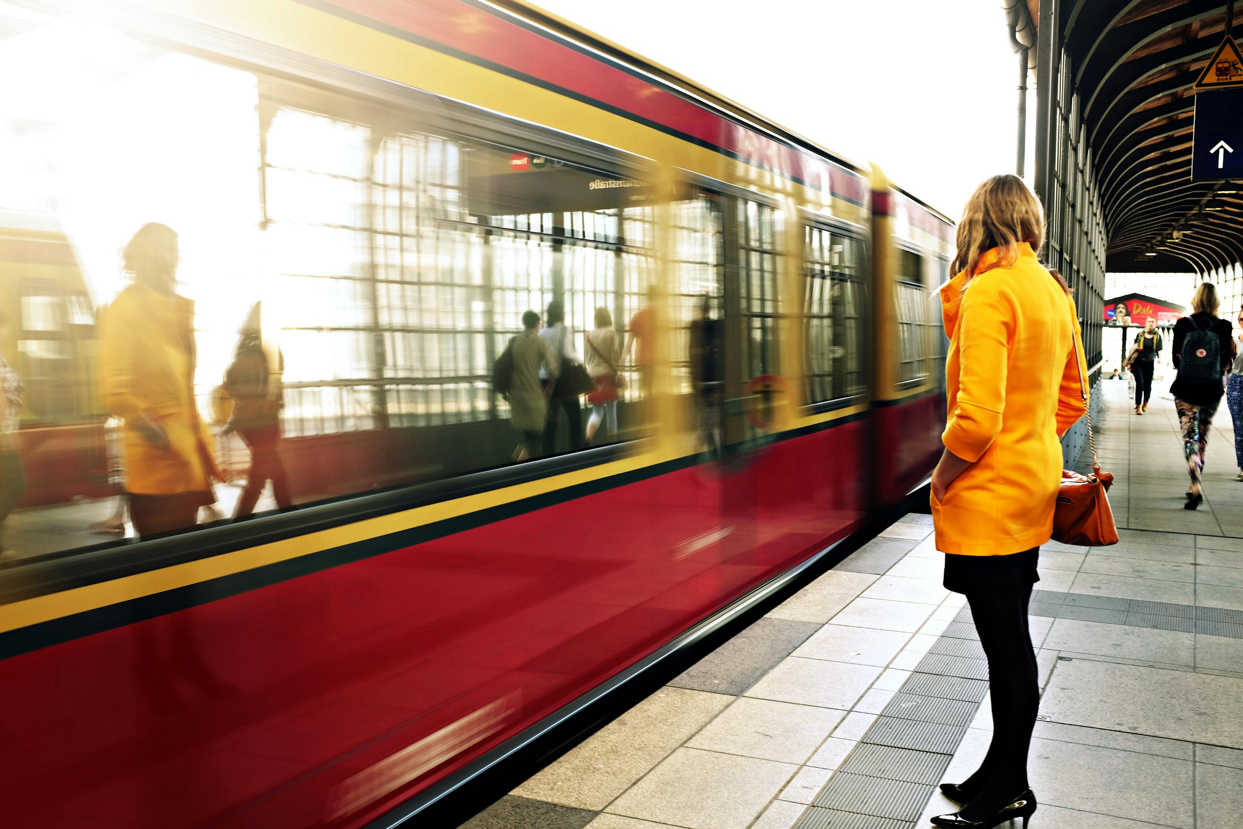 Woman in yellow coat waiting as red train passes in bustling station.