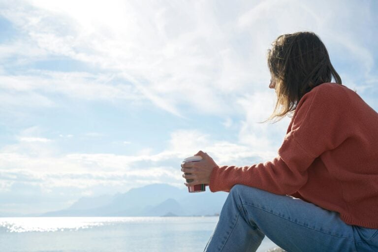 Woman in casual attire enjoys hot drink while gazing at the sea, relaxing.