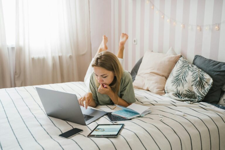 Woman lying on bed working remotely with laptop and tablet in bedroom