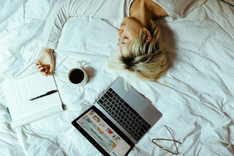 Woman sleeping with laptop, coffee, and notebook beside her in bed