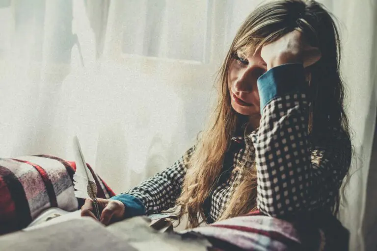 Woman in plaid shirt deep in thought by sunlit window with quill and paper.