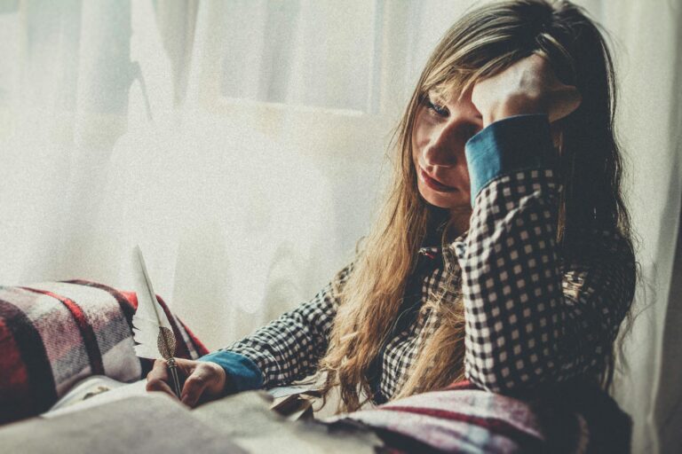 Woman in plaid shirt deep in thought by sunlit window with quill and paper.