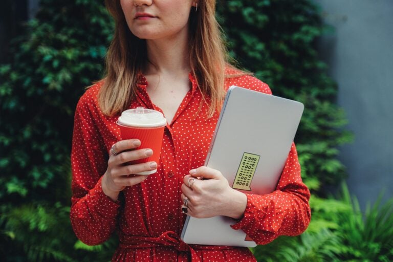 Young woman holding laptop and coffee cup outdoors wearing red polka dot dress.