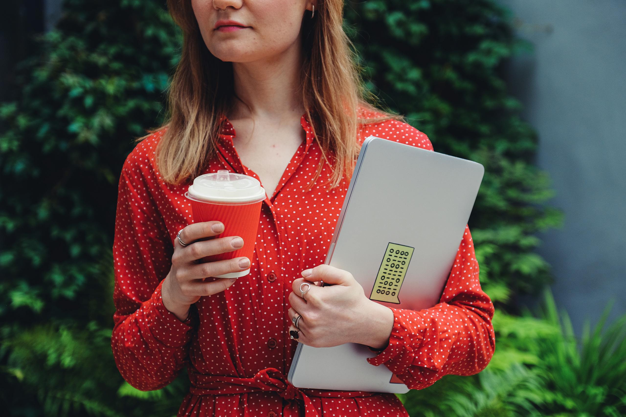 Young woman holding laptop and coffee cup outdoors wearing red polka dot dress.