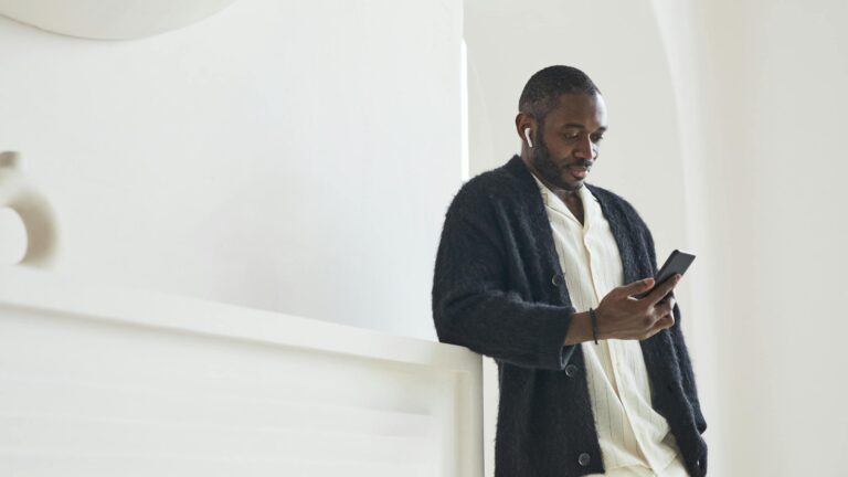 Fashionably dressed man using smartphone and earbuds indoors.